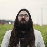 Colour photograph of Matthew E. White, who stands in front of a field of tall weeds; there are out-of-focus structures in the background, and the top half of the image is a light by overcast sky. White has long hair and a beard and wears glasses and a white suit-jacket with a colourful patterned shirt beneath and looks at the camera