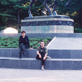 Band photograph of Evan Lewis and Tom Mcgreevy of Ducks Ltd., sat on the plinth of a statue of a horse, whose feet are seen at the top of the image.
