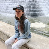 Colour photograph of Grouper, aka Liz Harris, sat on the ledge of a fountain wearing a black baseball cap, light-blue denim shirt and white jeans