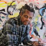 Colour photograph of Lonnie Holley, who leans, with glasses in hand, wearing a check-shirt, and looks at the camera; behind them is a colourful abstract painting