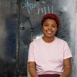 Photograph of Loraine James wearing a pink t-shirt, smiling and leaning against steel doors