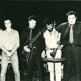 Black and white photograph of Pere Ubu. In the photograph, four members of the band stand somewhat awkwardly, posing with some of their instruments; they are mostly smiling