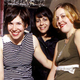 Candid band photograph of Carrie Brownstein, Janet Weiss, and Corin Tucker of Sleater-Kinney around 2002; the band are stood close together in a corridor and are looking at the camera smiling